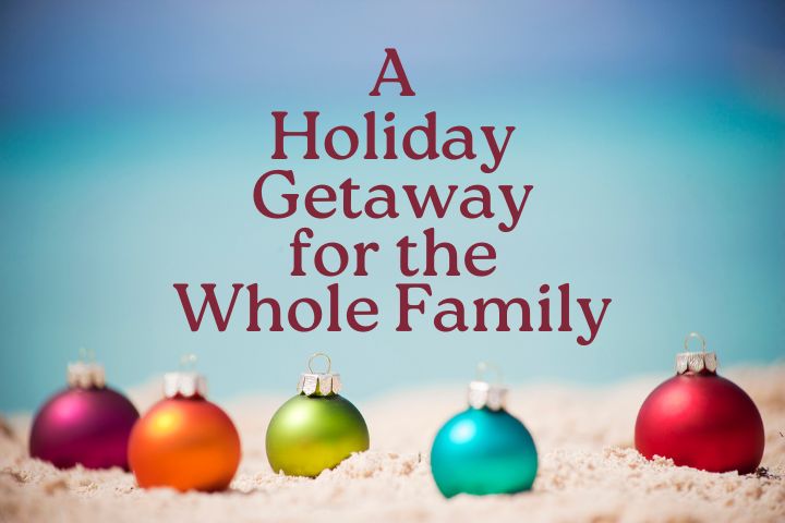 Five different colored ornaments placed in the sand with a sea blurred out in the back with the words in a maroon colors saying A Holiday Getaway for the Whole Family