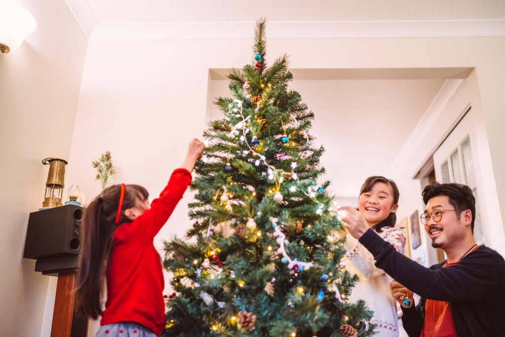 Young handsome dad hanging fairy lights on the Christmas tree with her two lovely daughters in living room at vacation rental.