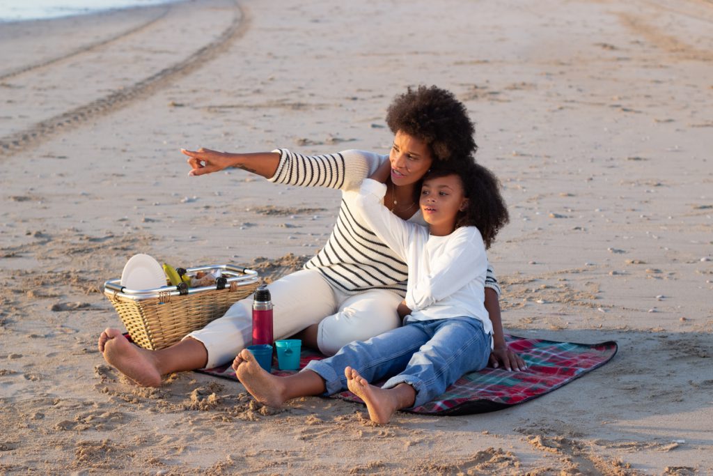 Relaxed mother and daughter having picnic on beach. African American mother and daughter in casual clothes sitting on blanket, discussing sunset. Family, relaxation, nature concept